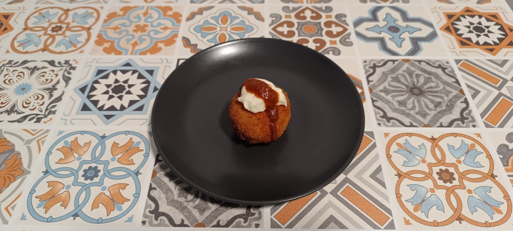 Crispy Bomba de la Barceloneta croquettes made from potatoes, topped with alioli and spicy red sauce, served on a black plate over Spanish tile table.
