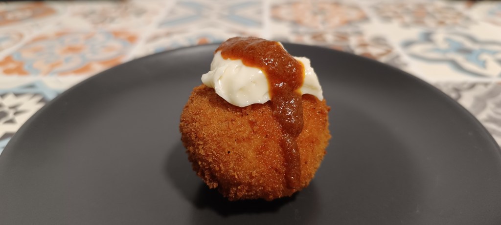 Golden potato Bomba de la Barceloneta croquettes with alioli and spicy red sauce, set on a black plate on a table with traditional Spanish tiles.
