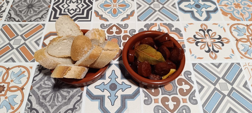 Juicy slices of chorizo in a light white wine sauce, served in a rustic tapas dish with pieces of bread on the side, all set on a colorful Spanish tiled table.