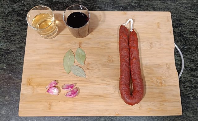 Flat lay of ingredients for chorizo al vino blanco: sliced Spanish chorizo, a bottle of white wine, garlic cloves, olive oil, fresh parsley, and crusty bread, all arranged on a decorative Spanish tile surface.