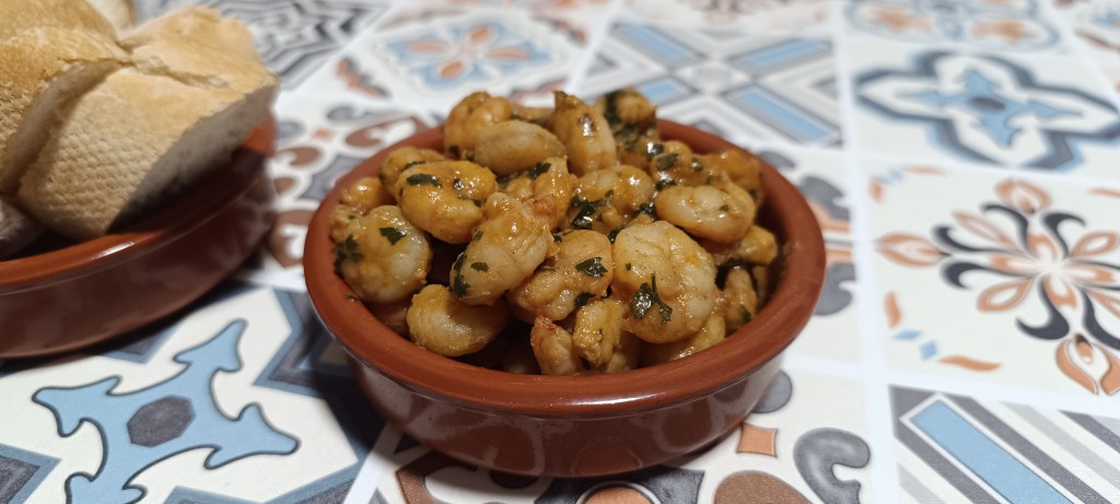A rustic tapas bowl filled with sizzling gambas al ajillo—garlicky prawns in olive oil with chili flakes—served with crusty bread on a colorful Spanish tiled table.