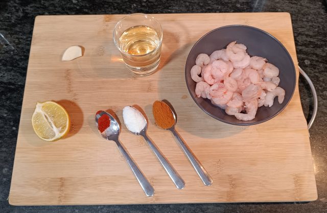 Flat lay of fresh ingredients for gambas al ajillo: raw prawns, sliced garlic, dried chili flakes, olive oil, parsley, and a loaf of crusty bread, arranged on a decorative Spanish tile background.