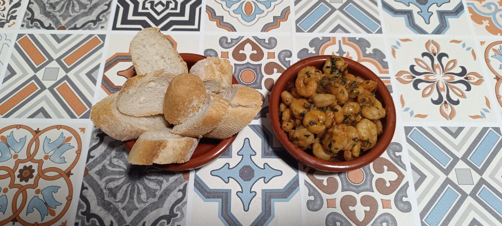 Gambas al ajillo served in a traditional clay tapas dish, with golden prawns swimming in garlic-infused olive oil, accompanied by slices of bread on a vibrant Spanish tile tabletop.