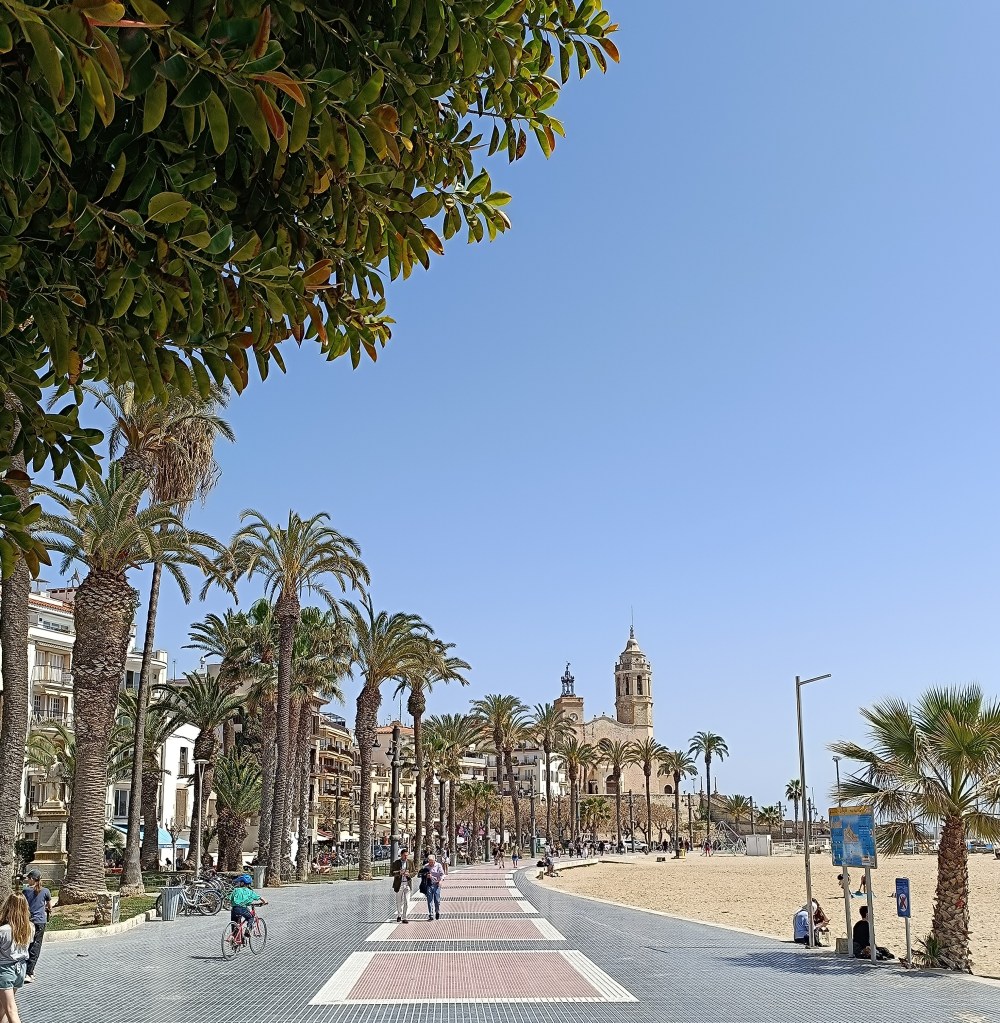 Historic church perched above the beach in Sitges, Spain, with waves lapping against the rocky shoreline.