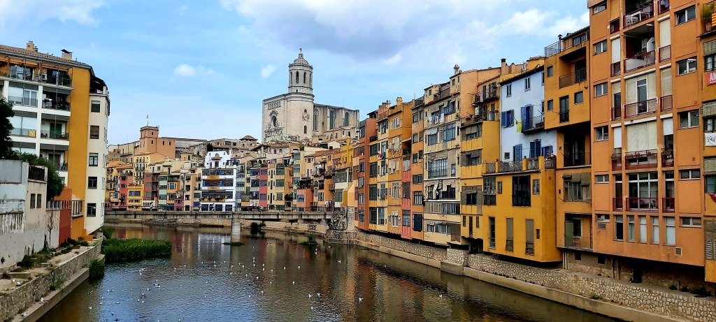 View of the Onyar River in Girona with a bridge crossing it, colorful buildings along the riverbanks, and the old town towers rising in the background