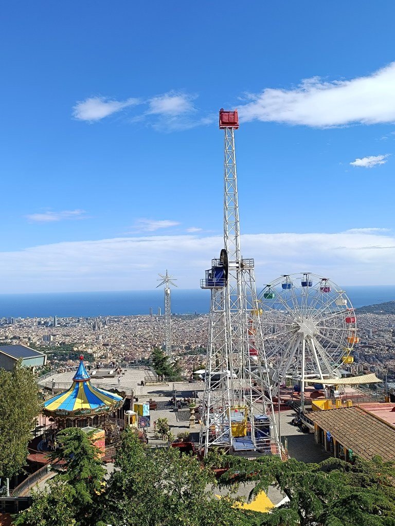 Panoramic view from Tibidabo showing the colorful amusement park rides in the foreground and Barcelona’s coastline with the Mediterranean Sea in the background.
