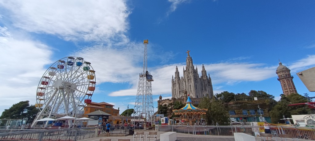View of Tibidabo Amusement Park with its colorful Ferris wheel in the foreground, and the Temple of the Sacred Heart church crowned by the statue of Jesus rising in the background