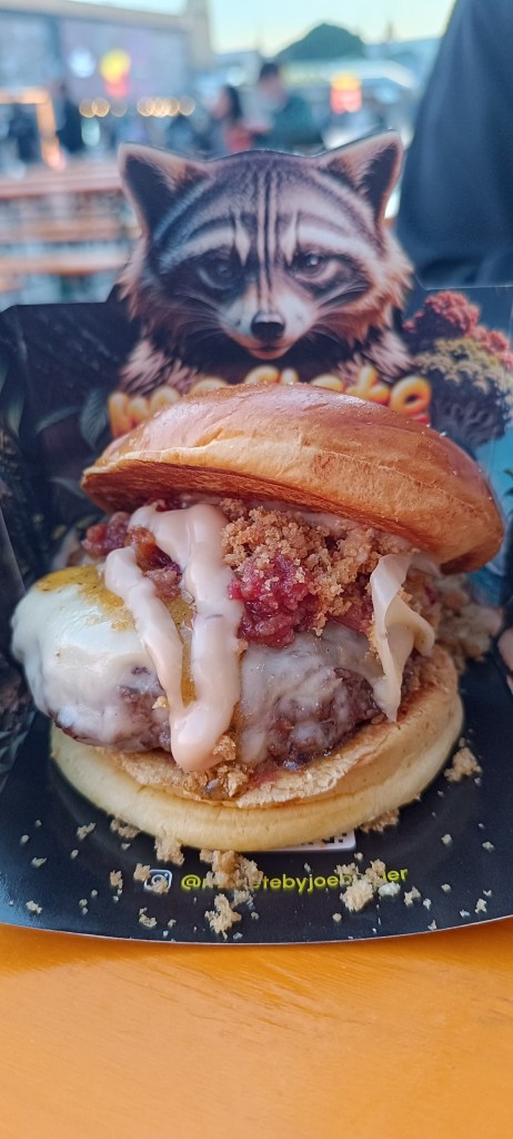 Burger stand with a raccoon logo at Champions Burger festival, surrounded by people waiting in line.