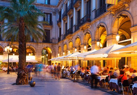 Plaça Reial in Barcelona at night, lit by streetlights, with palm trees, lively cafés, and people enjoying the evening atmosphere.